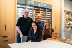 Four team members from Merit Kitchens smiling inside the design centre, with cabinet door samples and a display of wood finishes in the background.